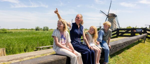 Drie kinderen krijgen uitleg van een molenaar in Kinderdijk met op de achtergrond een molen