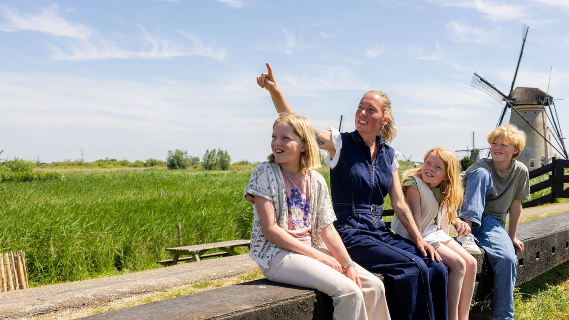 Drie kinderen krijgen uitleg van een molenaar in Kinderdijk met op de achtergrond een molen