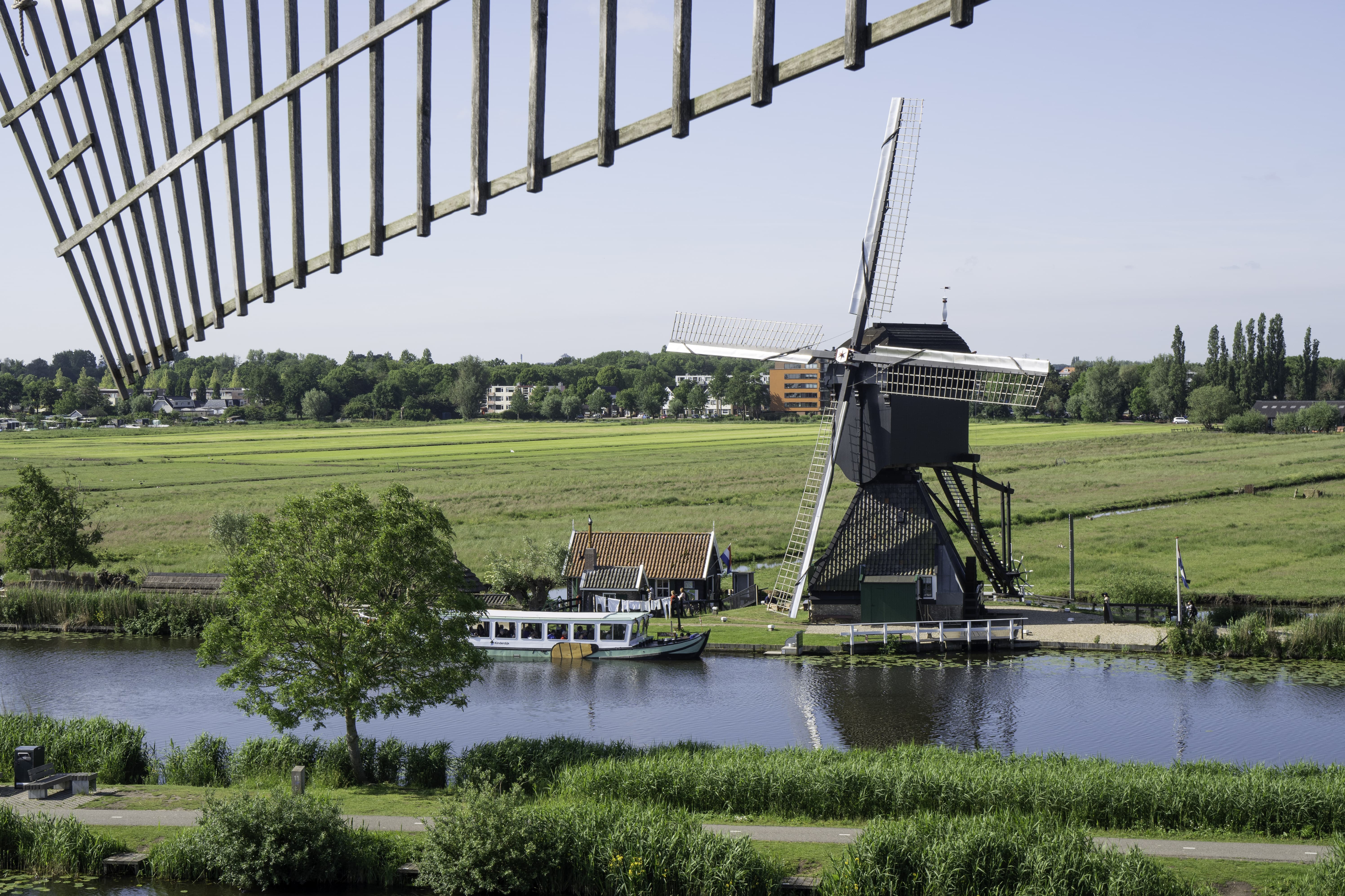 Photo taken from another windmill, with part of its sail still visible in the frame, looking toward the Blokweer Museum Mill in Kinderdijk. Behind it lies the polder, and in front of the mill there is a sightseeing boat.