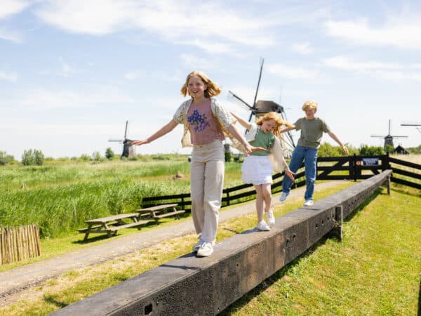 Drie kinderen spelen op de molenroede op het erf van Museummolen Nederwaard met op de achtergrond de molens van Kinderdijk