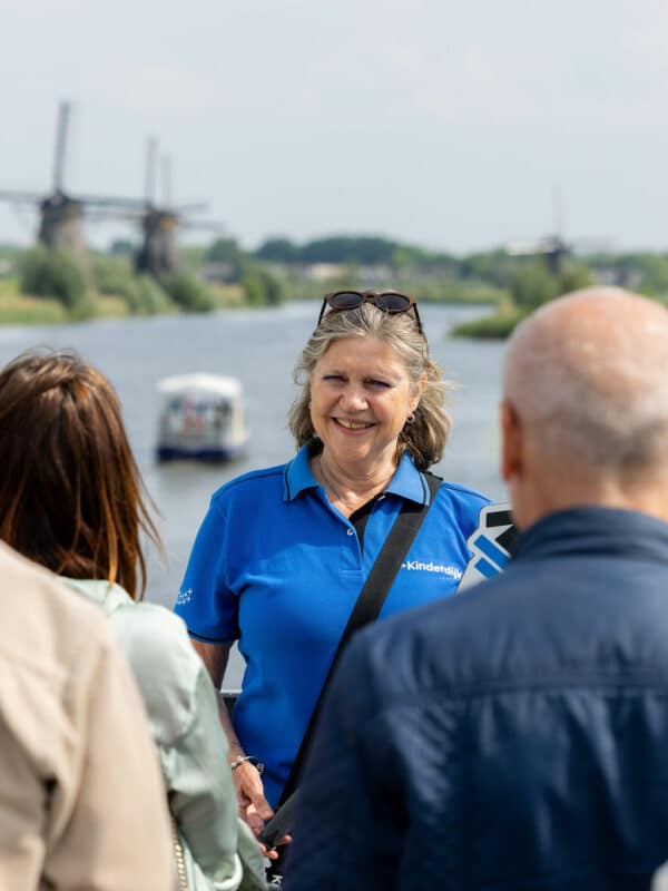 Een close-up van een vrijwillig gids van Kinderdijk. Ze heeft een groep voor zich en geeft hen uitleg, met op de achtergrond de rondvaartboot en molen.