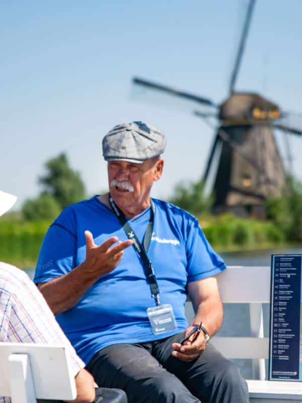 Een vrijwillige host geeft aan boord van de rondvaartboot uitleg aan bezoekers in Kinderdijk met de molens op de achtergrond.