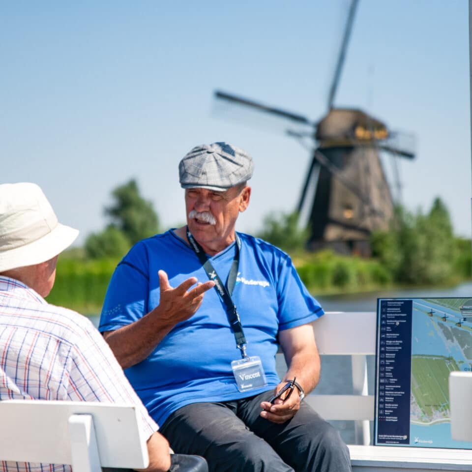 Een vrijwillige host geeft aan boord van de rondvaartboot uitleg aan bezoekers in Kinderdijk met de molens op de achtergrond.